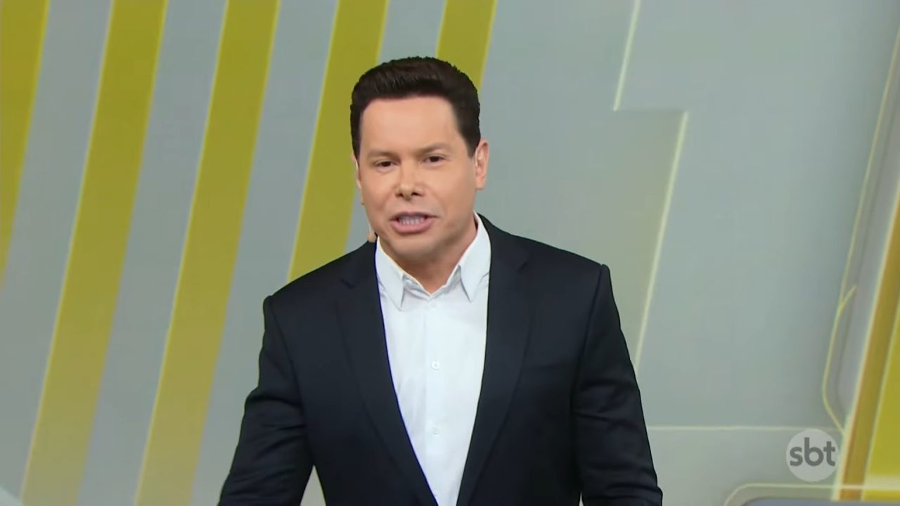 Marcão do Povo, apresentador do Primeiro Impacto, veste terno preto e fala no estúdio com fundo de linhas amarelas e logotipo da emissora.