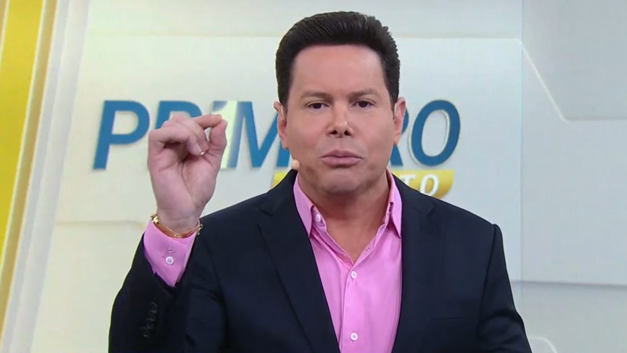 Marcão do Povo, apresentador do Primeiro Impacto, gesticula durante comentário ao vivo no estúdio do SBT com logotipo do jornal ao fundo.