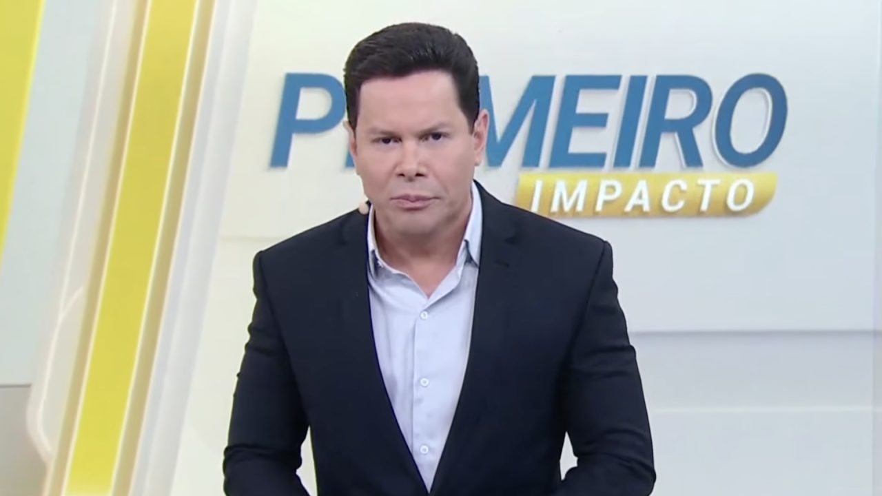 Marcão do Povo, de blazer escuro e camisa azul, encara a câmera no estúdio do Primeiro Impacto com logotipo do programa ao fundo