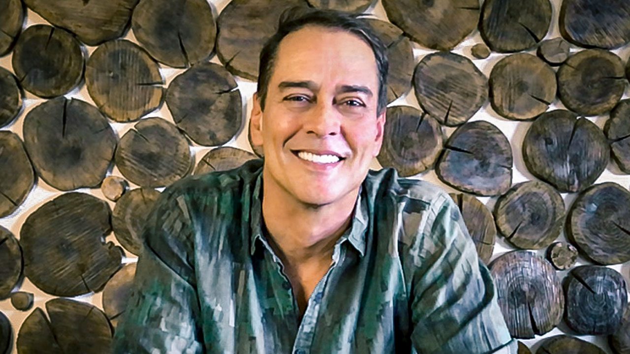 Marcello Antony sorrindo com camisa verde estampada diante de parede decorada com cortes circulares de madeira