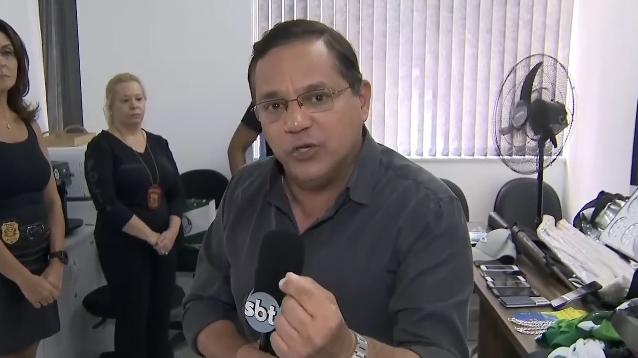 Repórter Marcelo Bittencourt segura microfone do SBT em delegacia durante reportagem com policiais ao fundo