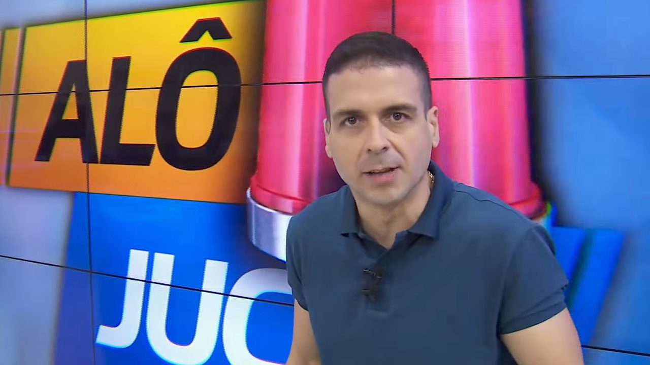 Marcelo Castro com camisa azul escura e colar, olhando para a câmera, com o logo do programa Alô Juca ao fundo