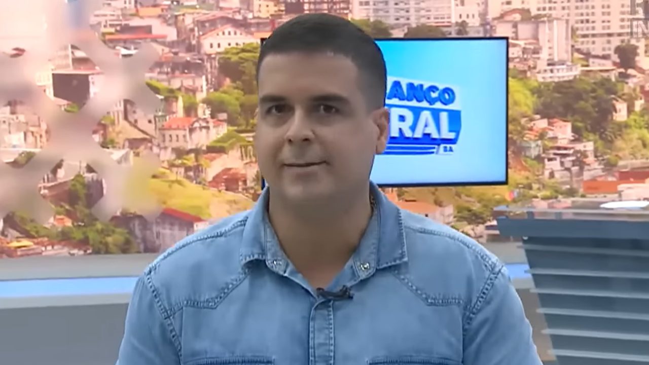 A imagem mostra o apresentador Marcelo Castro, do jornal Alô Juca, usando uma camisa jeans clara e microfone preso à gola. Ao fundo, há um painel com imagem panorâmica de uma cidade e uma tela exibindo o logotipo do programa Balanço Geral BA, em reportagem do Domingo Espetacular. O ambiente é bem iluminado e moderno, característico de um noticiário televisivo