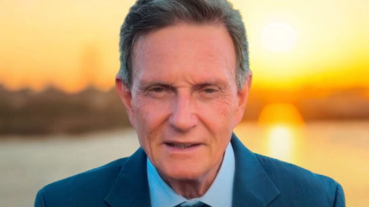 Marcelo Crivella de terno escuro com pôr do sol ao fundo em cenário ao ar livre