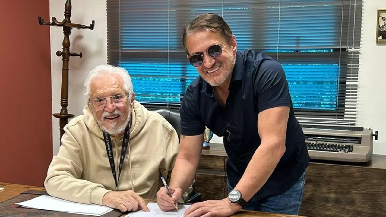 Homem idoso assina documento enquanto outro homem sorri ao lado em escritório durante encontro profissional