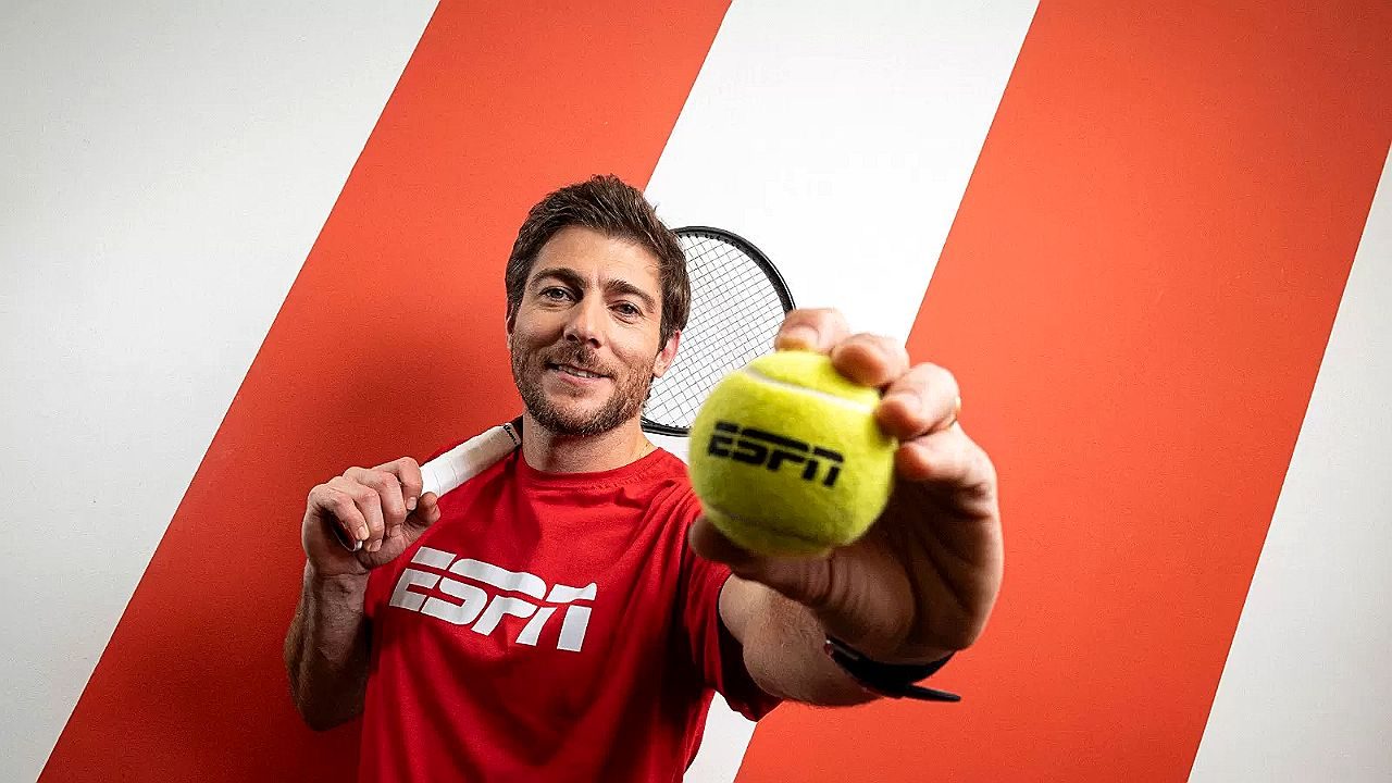 Homem segura bola de tênis com logo da ESPN diante de parede listrada e raquete nas costas