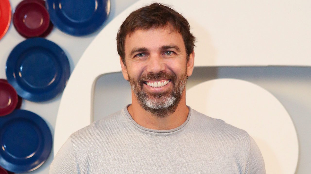 O ator Marcelo Faria com barba grisalha e camiseta cinza sorri para a câmera em frente a um painel decorado com pratos azuis e vermelhos