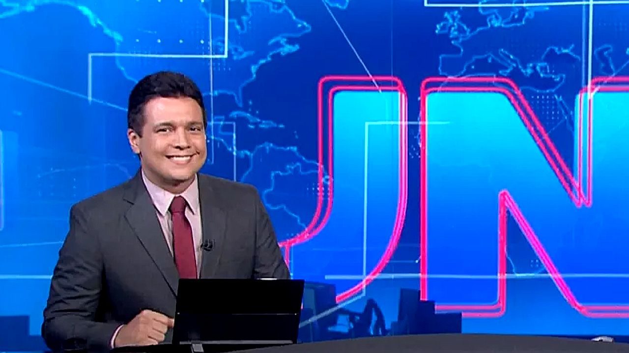 Apresentador de telejornal sorri atrás de bancada com notebook em estúdio com telão azul e sigla JN ao fundo