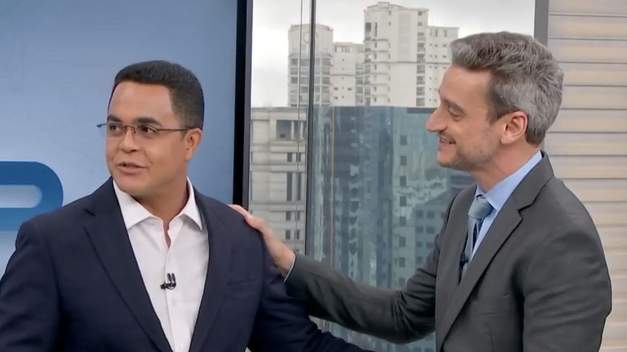Marcelo Pereira e Tiago Scheuer usam ternos e sorriem durante conversa no estúdio do telejornal Bom Dia São Paulo com vista de prédios ao fundo