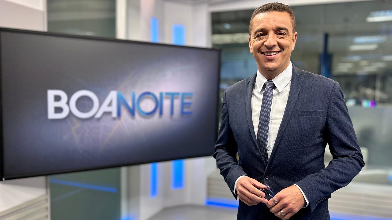 Marcelo Rocha de terno escuro sorri no estúdio do Boa Noite Paraná da RPC com o logo do programa na tela