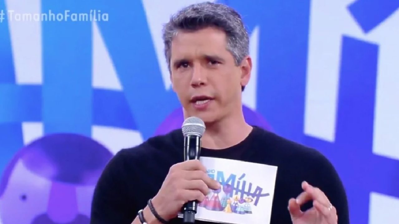 A imagem mostra um apresentador de televisão falando ao microfone em um estúdio colorido. Ele segura um cartão com o logotipo do programa “Tamanho Família” e aparece em frente a um fundo azul com o nome do programa em destaque