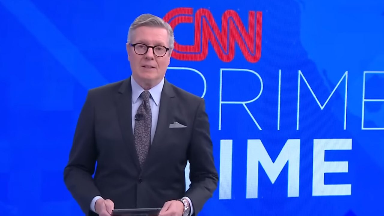 Márcio Gomes de terno escuro e óculos de grau apresenta o CNN Prime Time diante de telão azul com logotipo da CNN