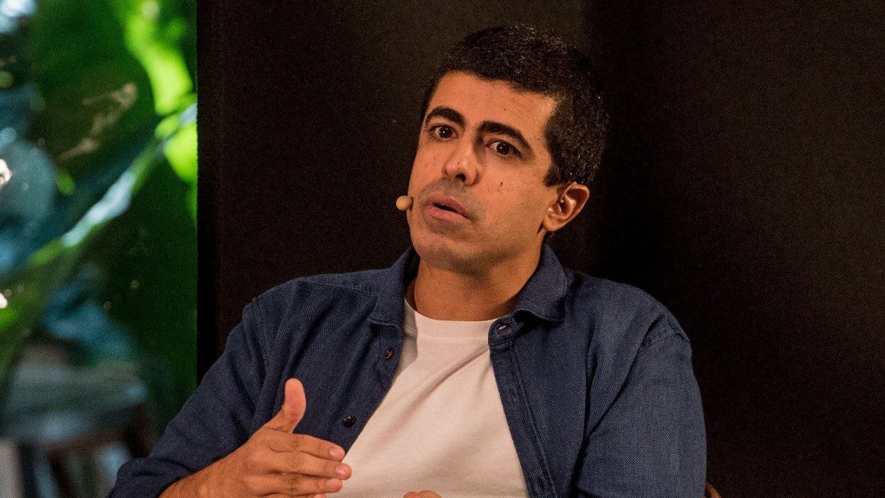 Marcius Melhem fala ao público com microfone de cabeça durante evento em ambiente interno com iluminação cênica ao fundo
