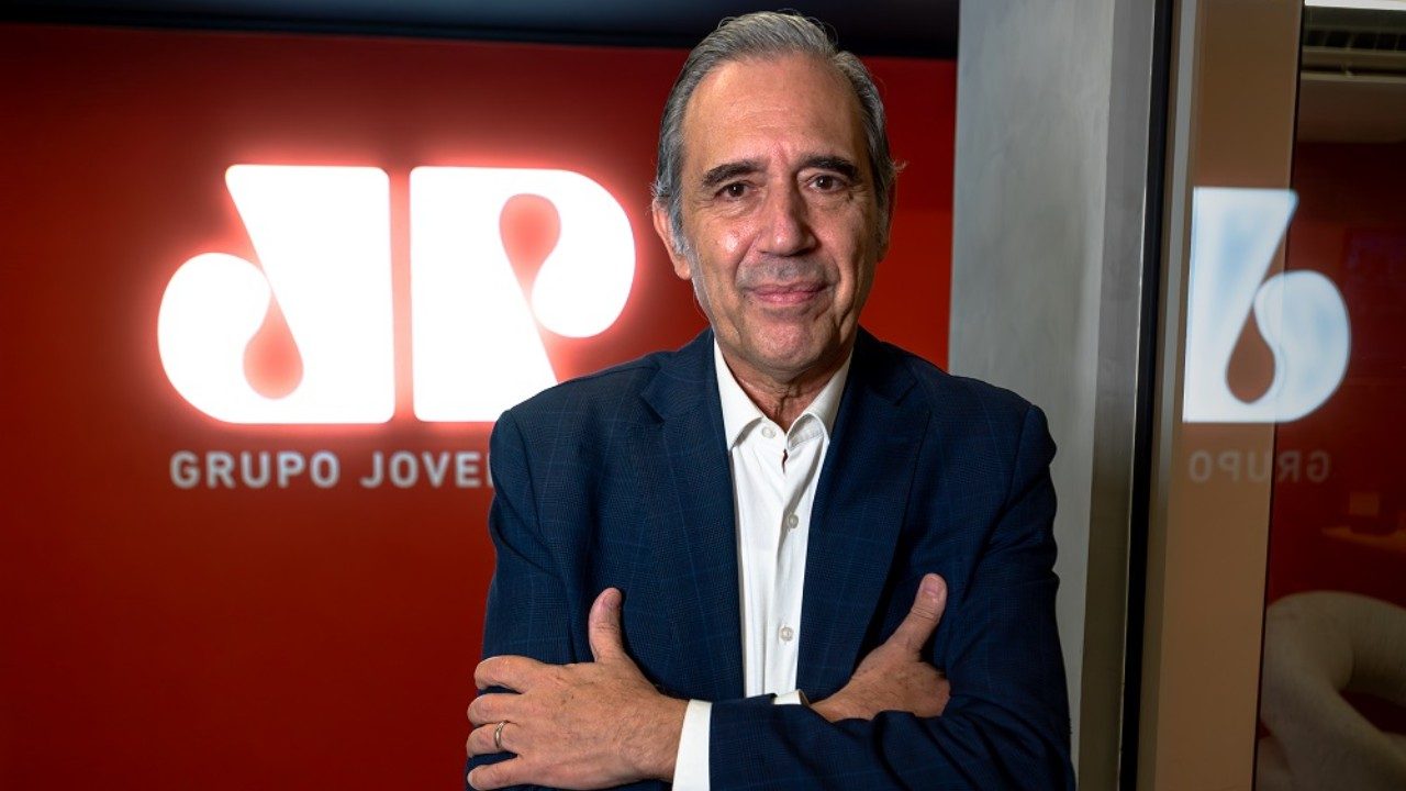 Marco Antonio Villa posa de braços cruzados em frente ao logotipo da Jovem Pan nos bastidores da emissora