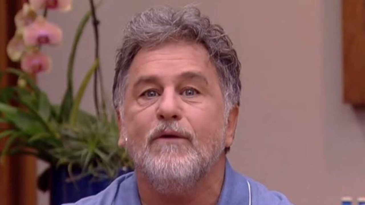 Homem de barba grisalha e camisa azul fala em ambiente interno com plantas ao fundo