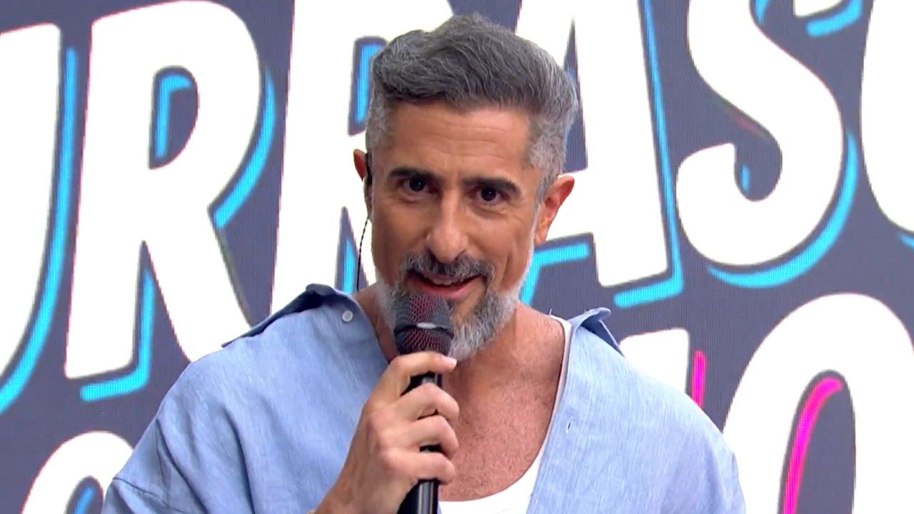 Marcos Mion segura microfone e sorri no palco do Caldeirão com Mion diante de painel com o nome do programa