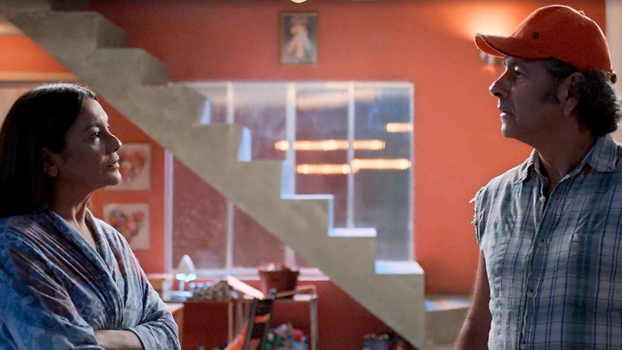 Homem de boné e camisa sem mangas conversa com mulher em sala com escada ao fundo em cena de novela