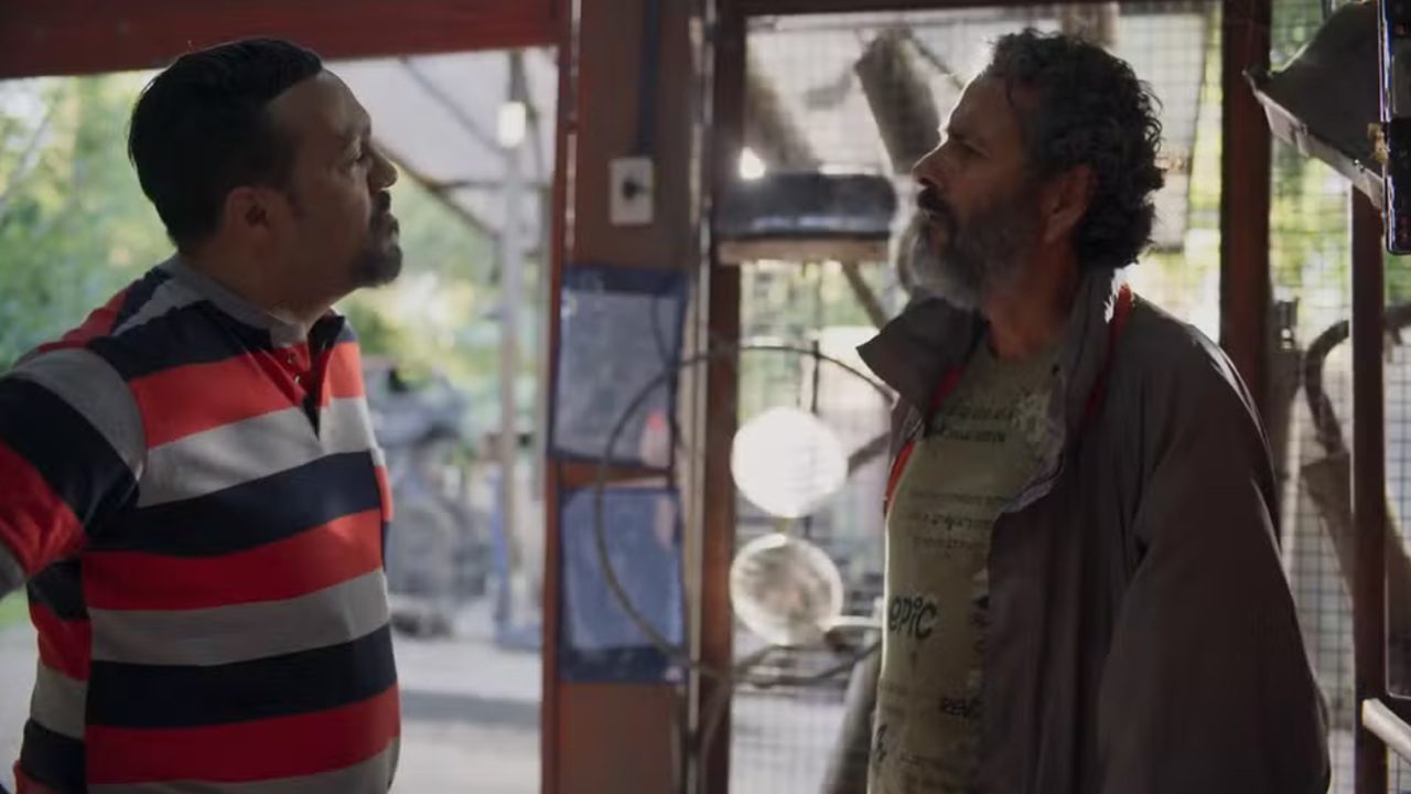 Dois homens conversam em um ambiente interno que lembra uma oficina ou galpão. À esquerda, um homem de cabelo curto e barba rala veste camisa polo listrada em vermelho, azul e branco e olha para o outro com expressão séria. À direita, um homem de cabelos grisalhos e barba cheia usa camiseta clara e jaqueta aberta, encarando o interlocutor. Ao fundo, há portas de vidro, ventiladores e estruturas metálicas, com luz natural entrando pelo ambiente.