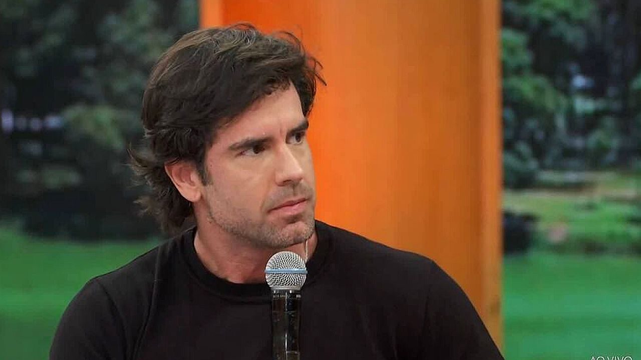 Homem de camiseta preta fala ao microfone durante programa de TV com cenário ao fundo