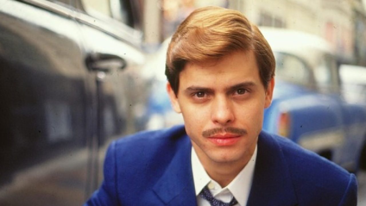 Homem jovem encara a câmera em um retrato ao ar livre. Ele tem cabelos claros bem penteados de lado, usa um bigode fino e veste terno azul com camisa clara e gravata. O fundo mostra carros estacionados em uma rua urbana, desfocados, criando profundidade e destacando o rosto e a expressão séria do personagem.