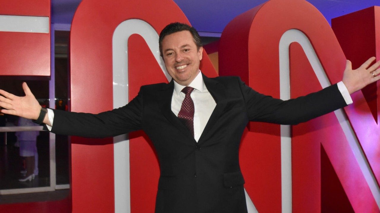 Marcus Chisco de terno e gravata sorri com braços abertos em frente ao logotipo da CNN em ambiente interno iluminado