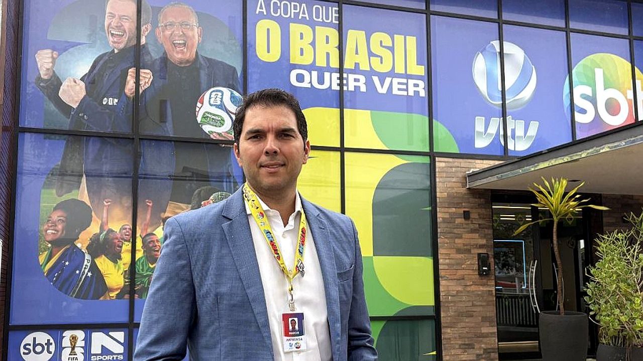 Homem com credencial posa em frente à sede do SBT com painel da Copa do Mundo e logos da emissora