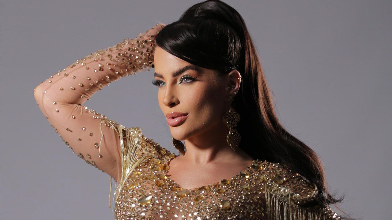 Mari Marquini com rabo de cavalo alto, usando vestido dourado com franjas e brilhos, posando com a mão na cabeça