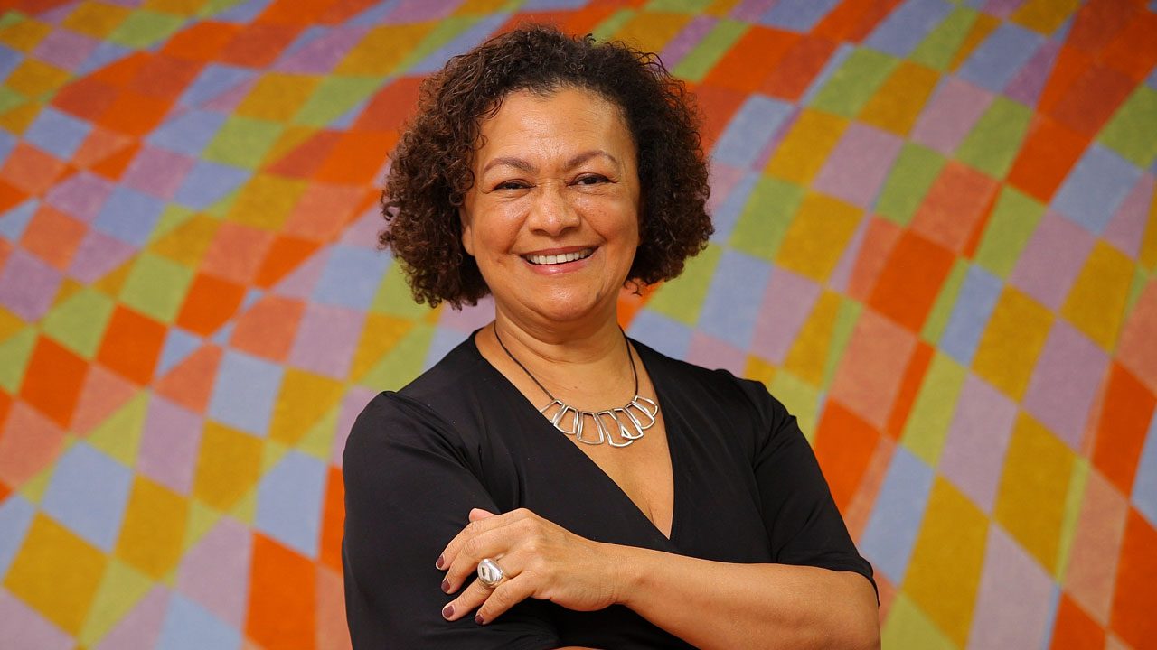 Maria Angela de Jesus, nova presidente da TV Cultura, sorri com os braços cruzados em frente a parede colorida com padrão geométrico