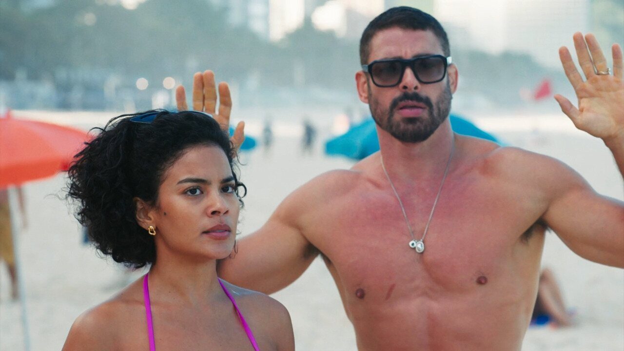 Maria de Fátima (Bella Campos) e César (Cauã Reymond) de mãos erguidas em cena na praia no remake de Vale Tudo