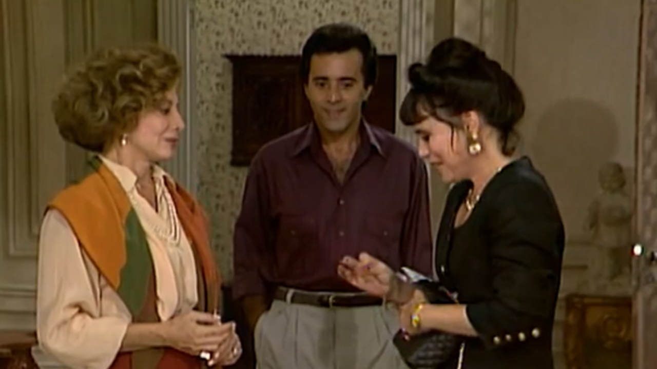 Cena mostra duas mulheres e um homem em uma sala decorada; as mulheres conversam cordialmente, enquanto o homem observa ao fundo sorrindo, em ambiente com tons clássicos e elegantes