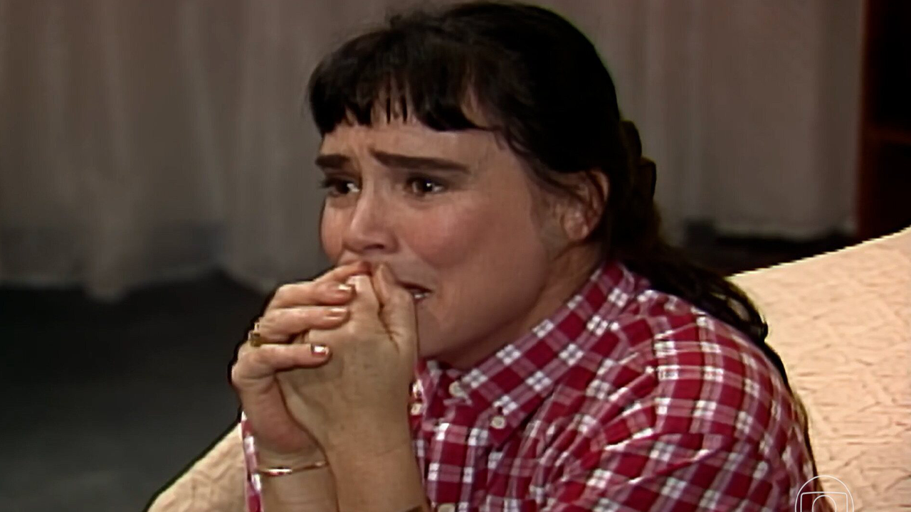 Regina Duarte como Maria do Carmo aparece aflita com mãos próximas ao rosto em cena da novela Rainha da Sucata em ambiente interno