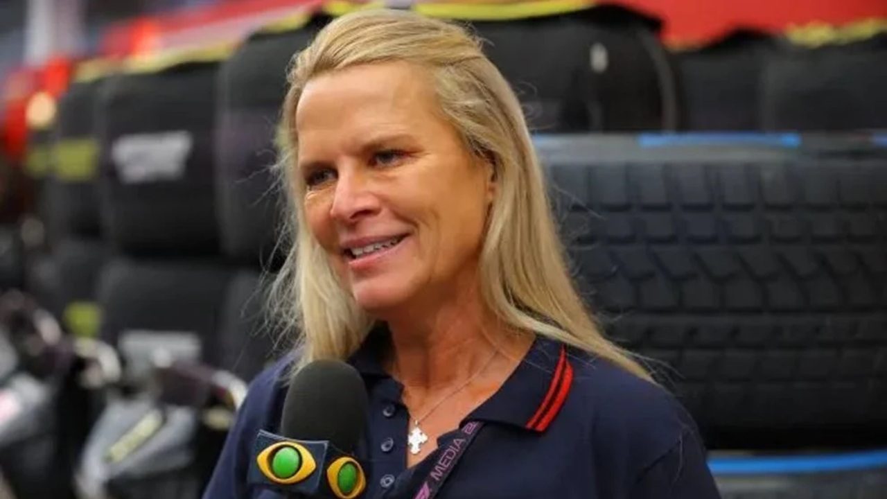 Mulher loira sorridente, segurando um microfone com o logo amarelo e verde de um canal de TV; ela veste camisa azul marinho com detalhes vermelhos, e ao fundo há pneus empilhados e equipamentos automotivos, indicando ambiente de cobertura esportiva.