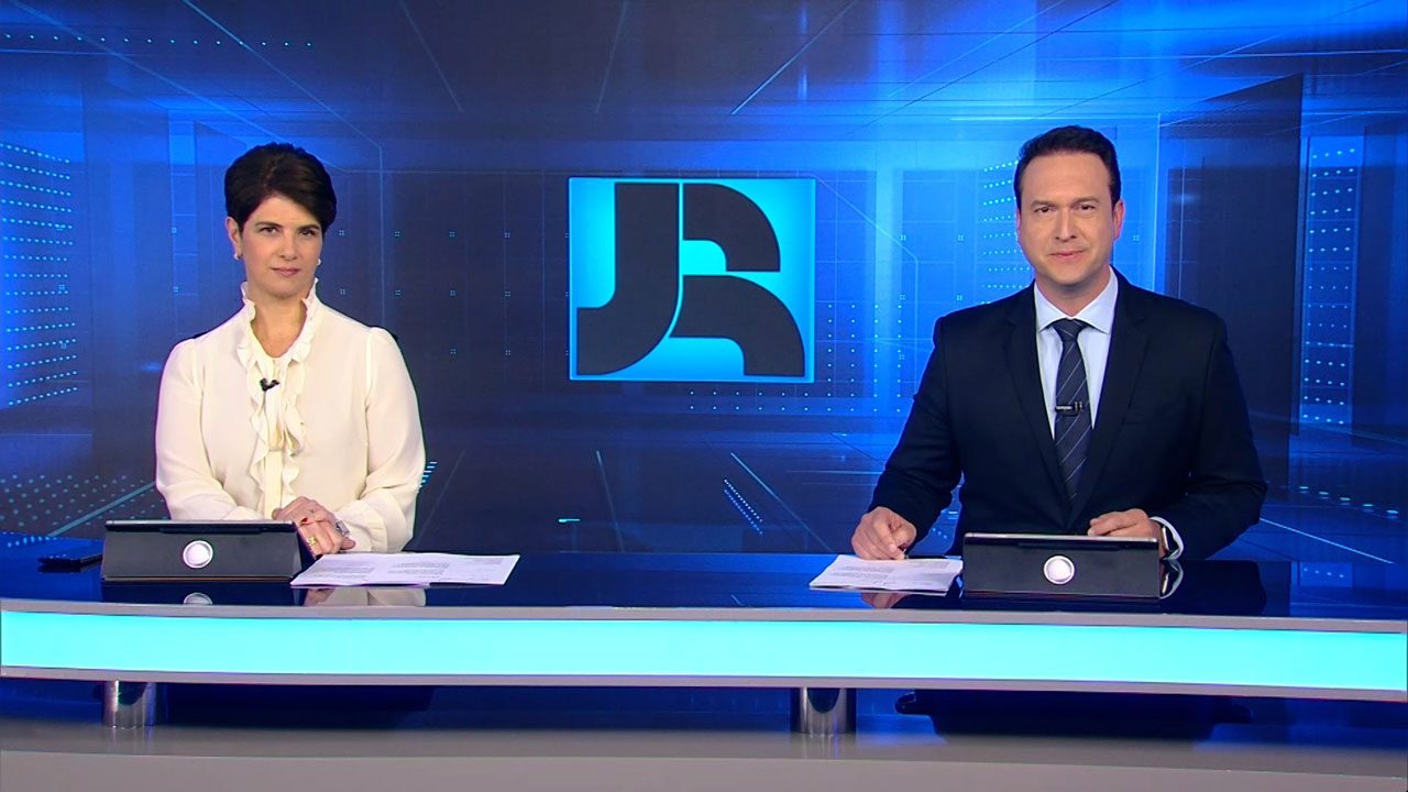 Mariana Godoy e Eduardo Ribeiro sentados na bancada do Jornal da Record com logotipo ao fundo