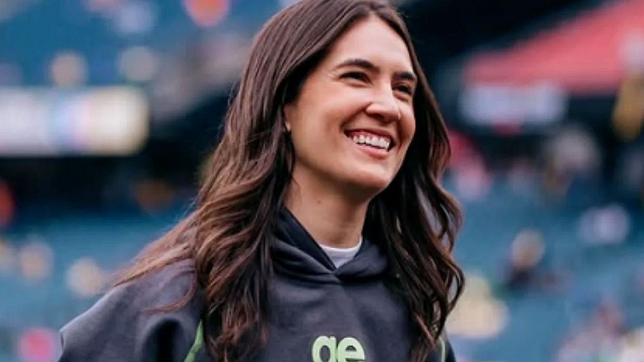 Mulher de cabelos castanhos longos sorri durante presença em estádio esportivo usando moletom escuro com arquibancadas desfocadas ao fundo