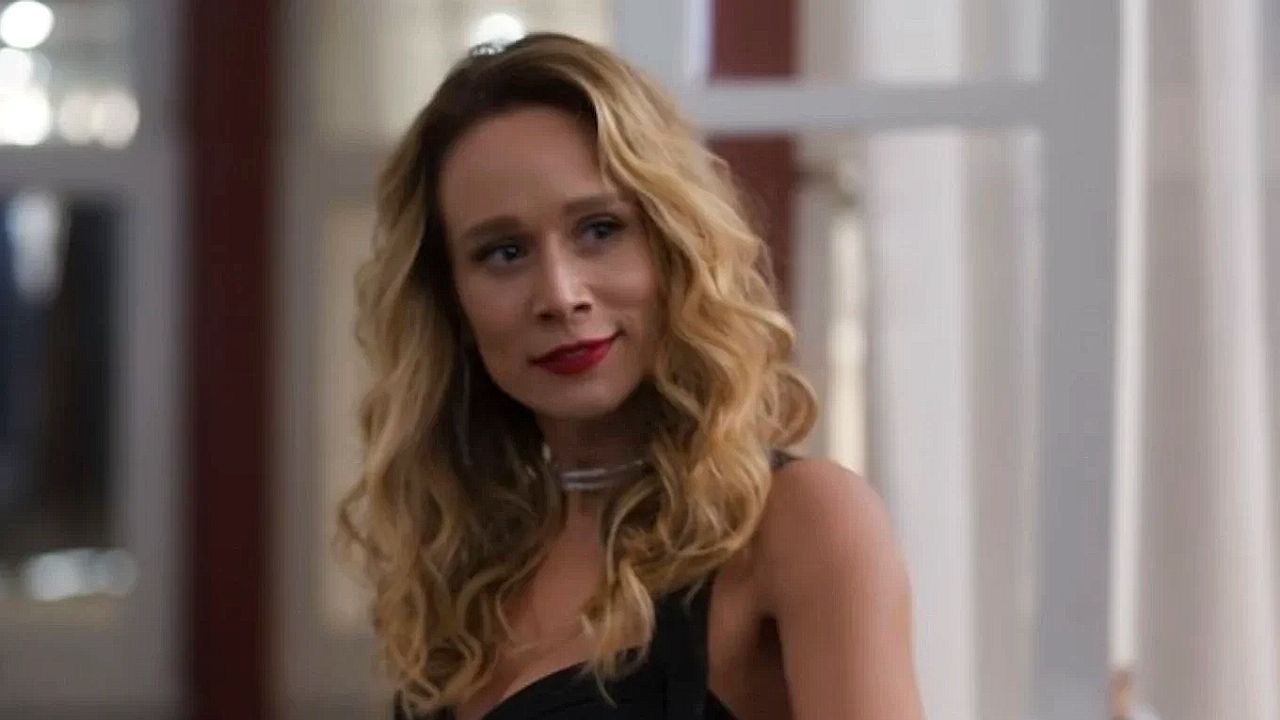 Atriz de cabelos loiros ondulados e vestido preto observa alguém com leve sorriso em cena de novela brasileira