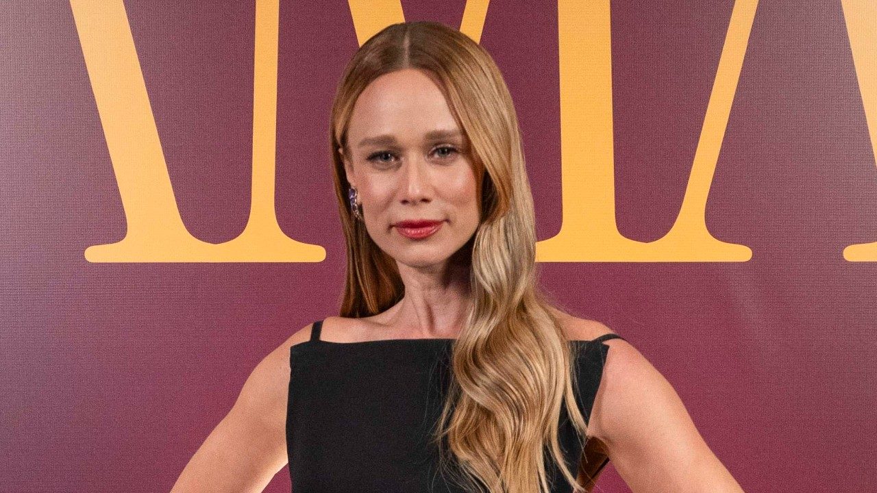 Mariana Ximenes posa com vestido preto e cabelo loiro ondulado em evento com fundo vinho e letras douradas