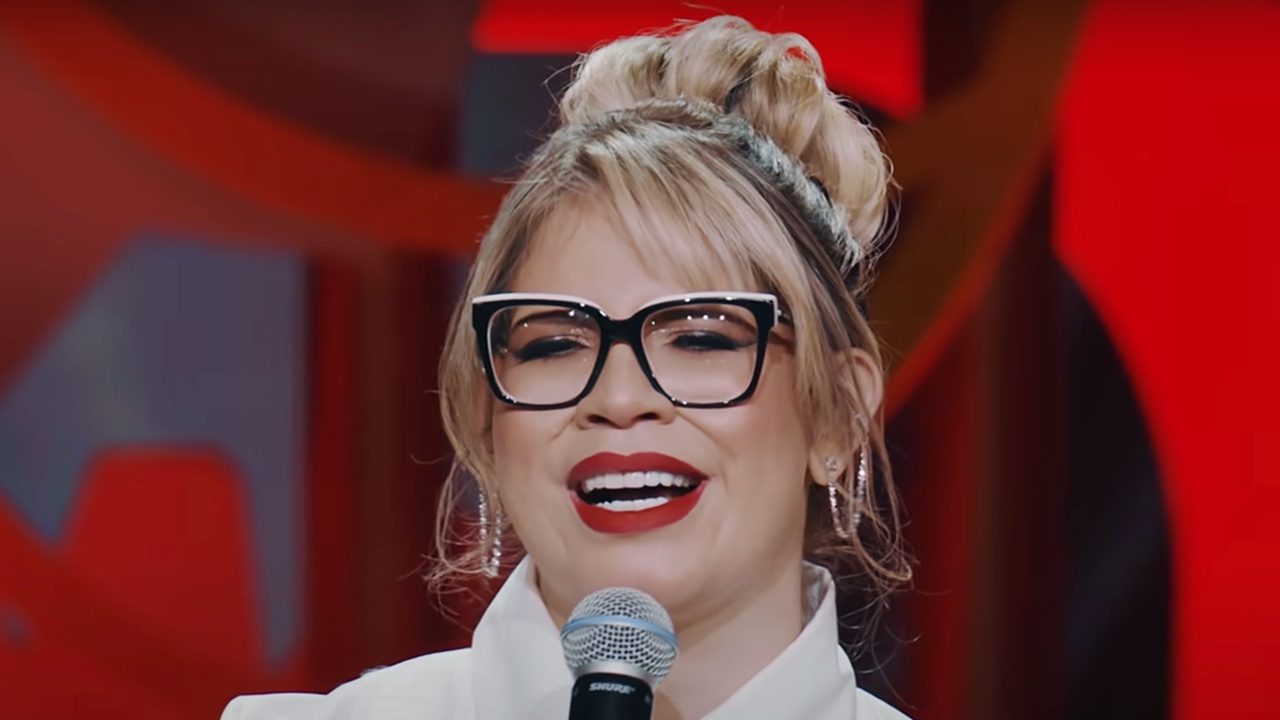 Mulher sorrindo enquanto segura um microfone em um estúdio iluminado por tons de vermelho. Ela usa óculos de armação grossa preta com detalhes claros, batom vermelho intenso e cabelo loiro preso em um coque alto com mechas soltas. A expressão é alegre e confiante, destacando o clima de apresentação ou performance ao vivo.