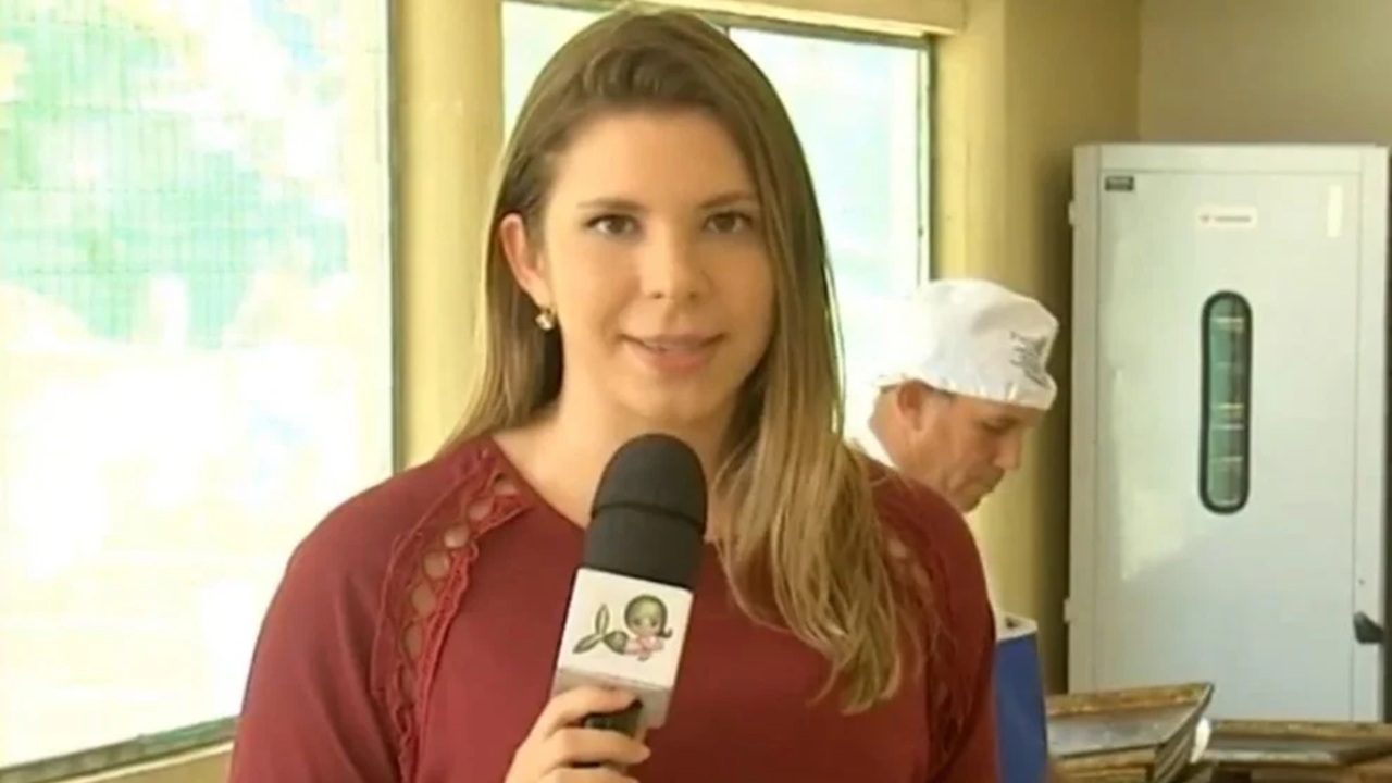 Repórter segura um microfone enquanto faz uma entrada ao vivo em um ambiente interno iluminado. Ela usa blusa vinho e está em primeiro plano, olhando para a câmera. Ao fundo, um profissional com touca branca trabalha próximo a uma bancada