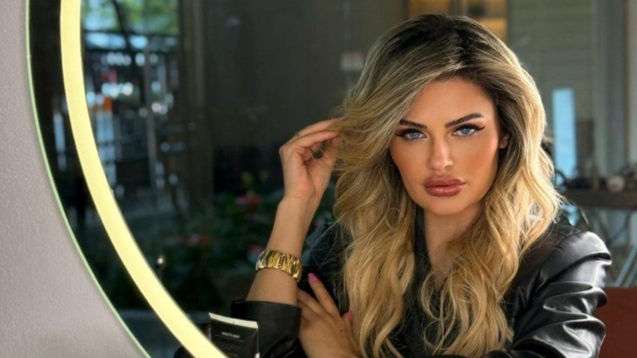 Marina Smith posa com olhar direto enquanto ajusta o cabelo diante de espelho iluminado em ambiente interno