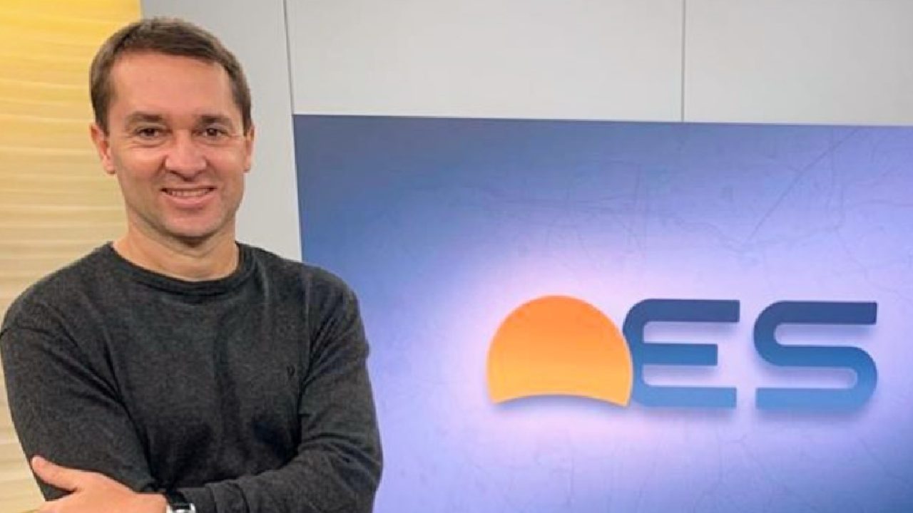 A imagem mostra um homem sorridente de braços cruzados, vestindo uma blusa escura, em um estúdio de televisão. Atrás dele, há um painel com o logotipo “ES”, que parece fazer parte de um programa jornalístico. O ambiente é bem iluminado e transmite uma atmosfera profissional