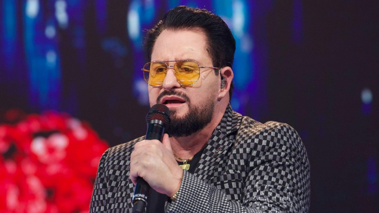 Marrone canta ao microfone com óculos amarelos e blazer estampado em apresentação no palco com luzes coloridas ao fundo