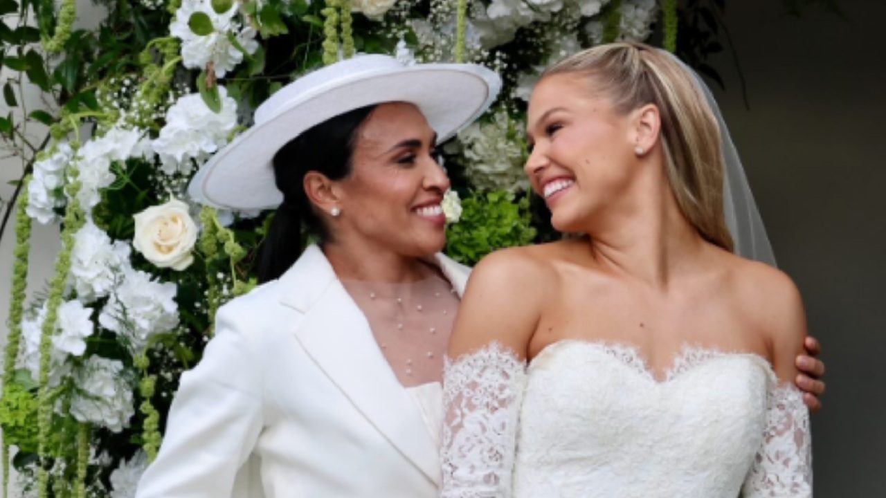 Duas mulheres sorriem uma para a outra em frente a um arco decorado com flores brancas e folhagens. A mulher à esquerda usa terno branco e chapéu combinando, enquanto a mulher à direita veste um vestido de noiva branco com renda e véu preso aos cabelos loiros.