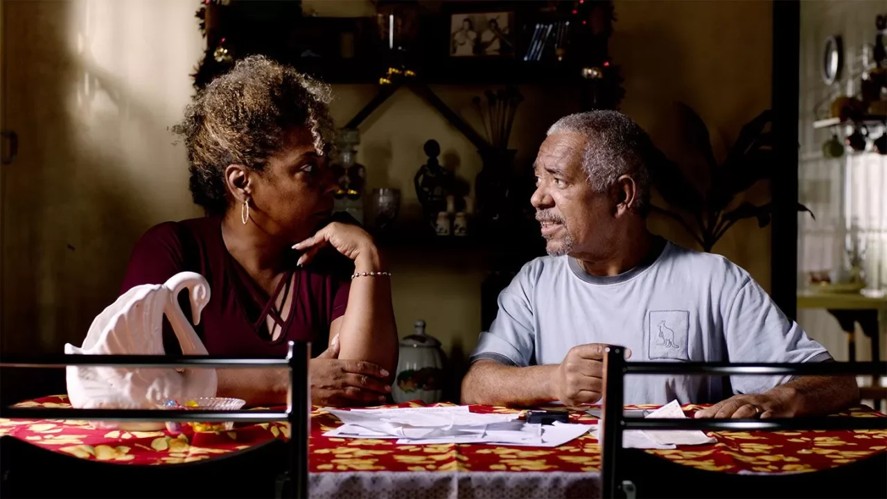Uma mulher e um homem conversando em uma sala escura com uma mesa de centro