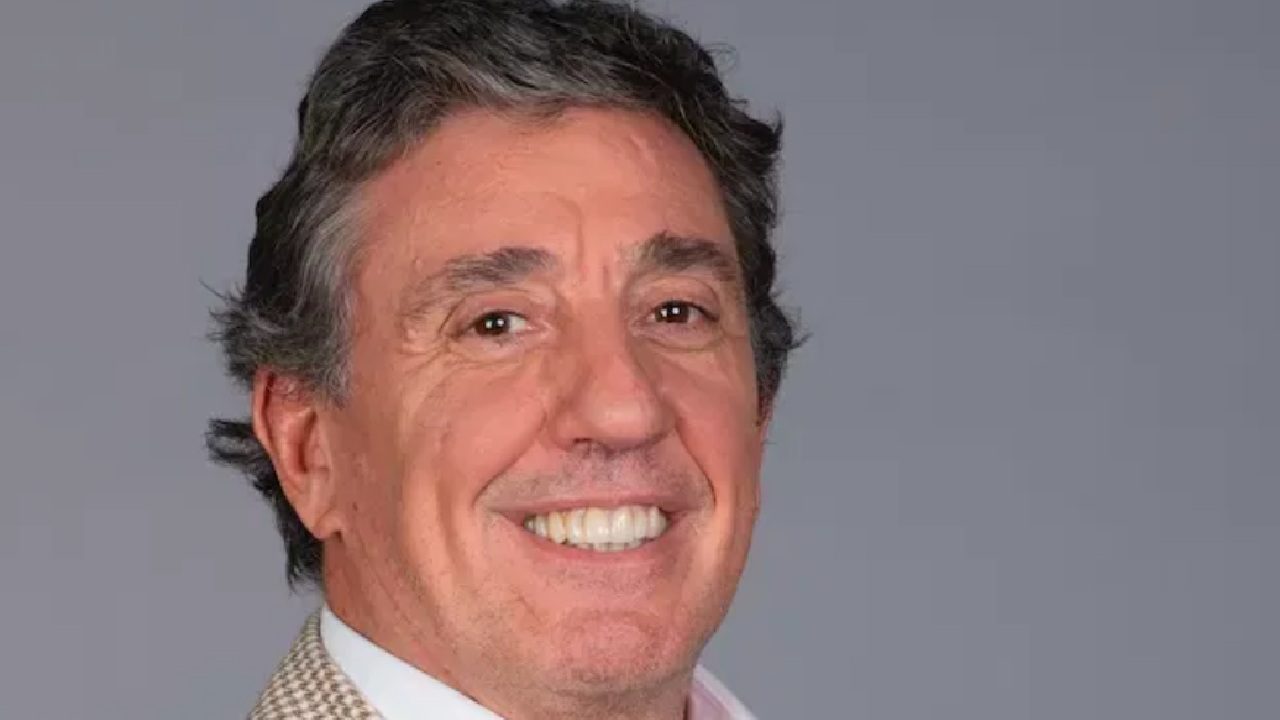 Homem de meia-idade sorri para a câmera em um retrato de estúdio, com fundo neutro cinza e traje social claro