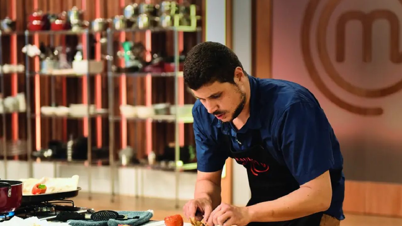 A imagem mostra um participante de um programa culinário, concentrado enquanto prepara um prato em uma bancada de cozinha. Ele usa um avental preto e camisa azul, e ao fundo há prateleiras com panelas e utensílios. O cenário tem o logotipo do programa “MasterChef” visível na parede