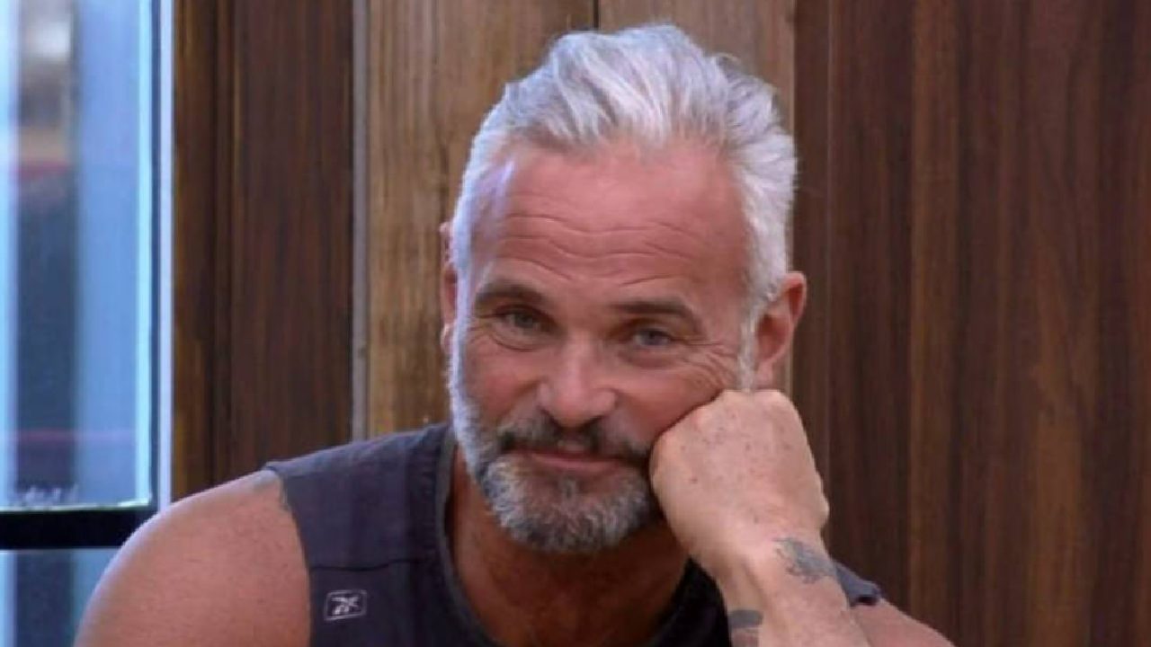 Homem de cabelos grisalhos e barba curta aparece em ambiente interno, apoiando o rosto na mão direita e sorrindo de forma discreta. Ele veste camiseta sem mangas escura, revelando parte de uma tatuagem no braço. Ao fundo, há uma parede de madeira e uma janela, compondo um cenário simples e iluminado.