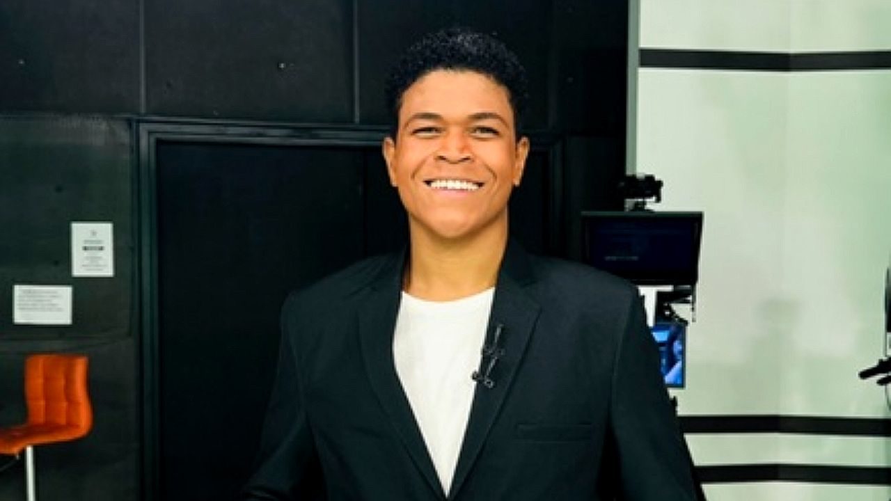Homem sorri nos bastidores de estúdio de TV usando blazer preto e microfone de lapela
