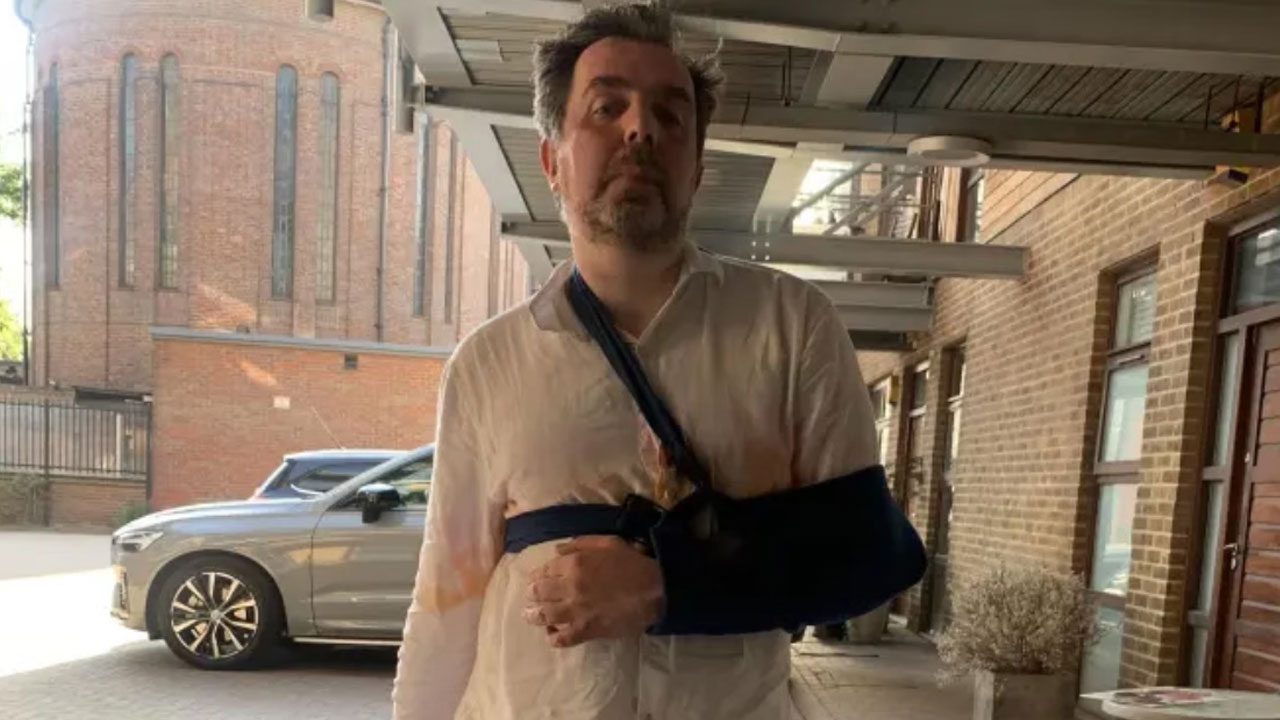 Homem de camisa branca com o braço esquerdo imobilizado em tipoia azul caminhando em área externa com prédio de tijolos ao fundo