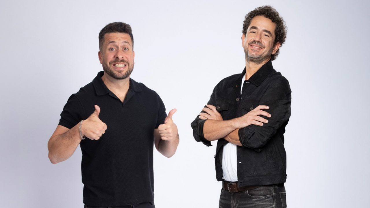 Dois apresentadores posam para foto promocional com roupas escuras e gestos descontraídos diante de fundo neutro
