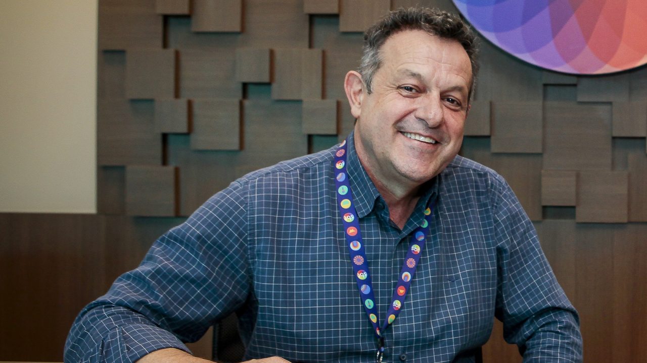 Mauro Lissoni, homem de meia-idade, sorri para a câmera em ambiente corporativo do SBT, usando camisa xadrez azul e crachá colorido no pescoço; ao fundo, parede com revestimento geométrico de madeira e parte de logotipo institucional